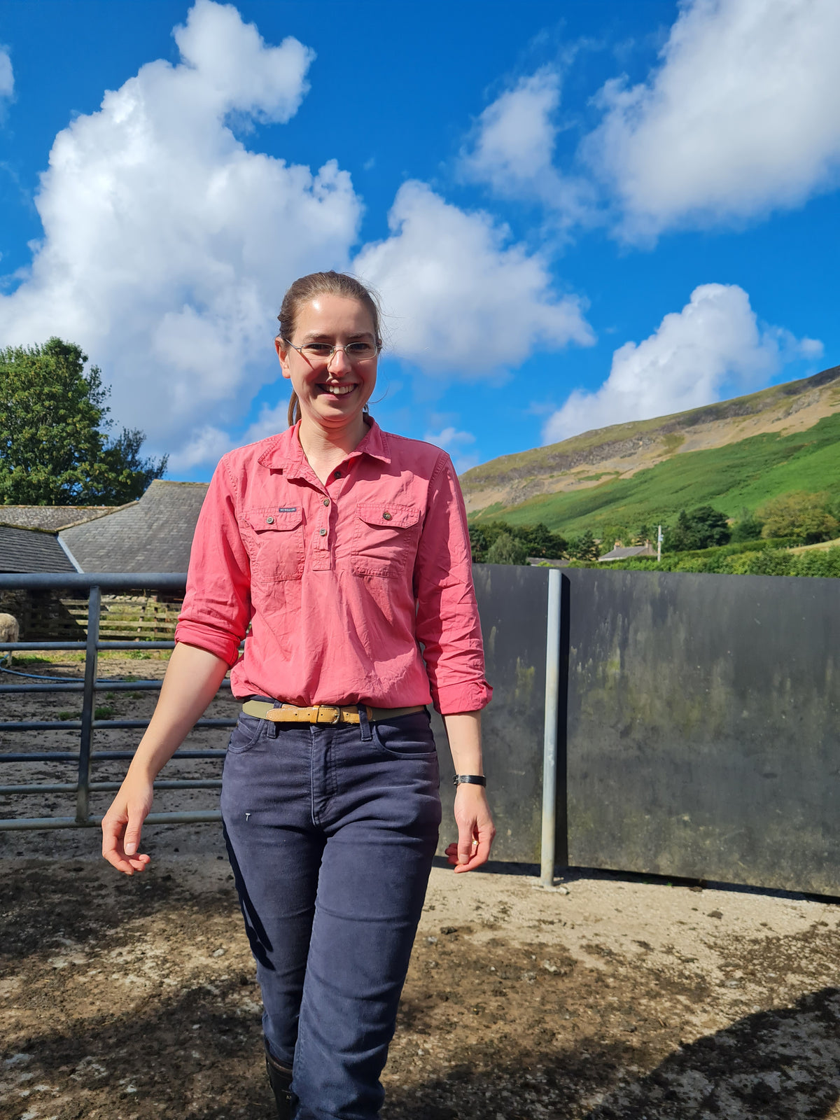 Isobel from Bleak Ridge Clothing wearing a pink button-up shirt and navy trousers stands outdoors next to a metal farm fence, with a green field and blue sky in the background.