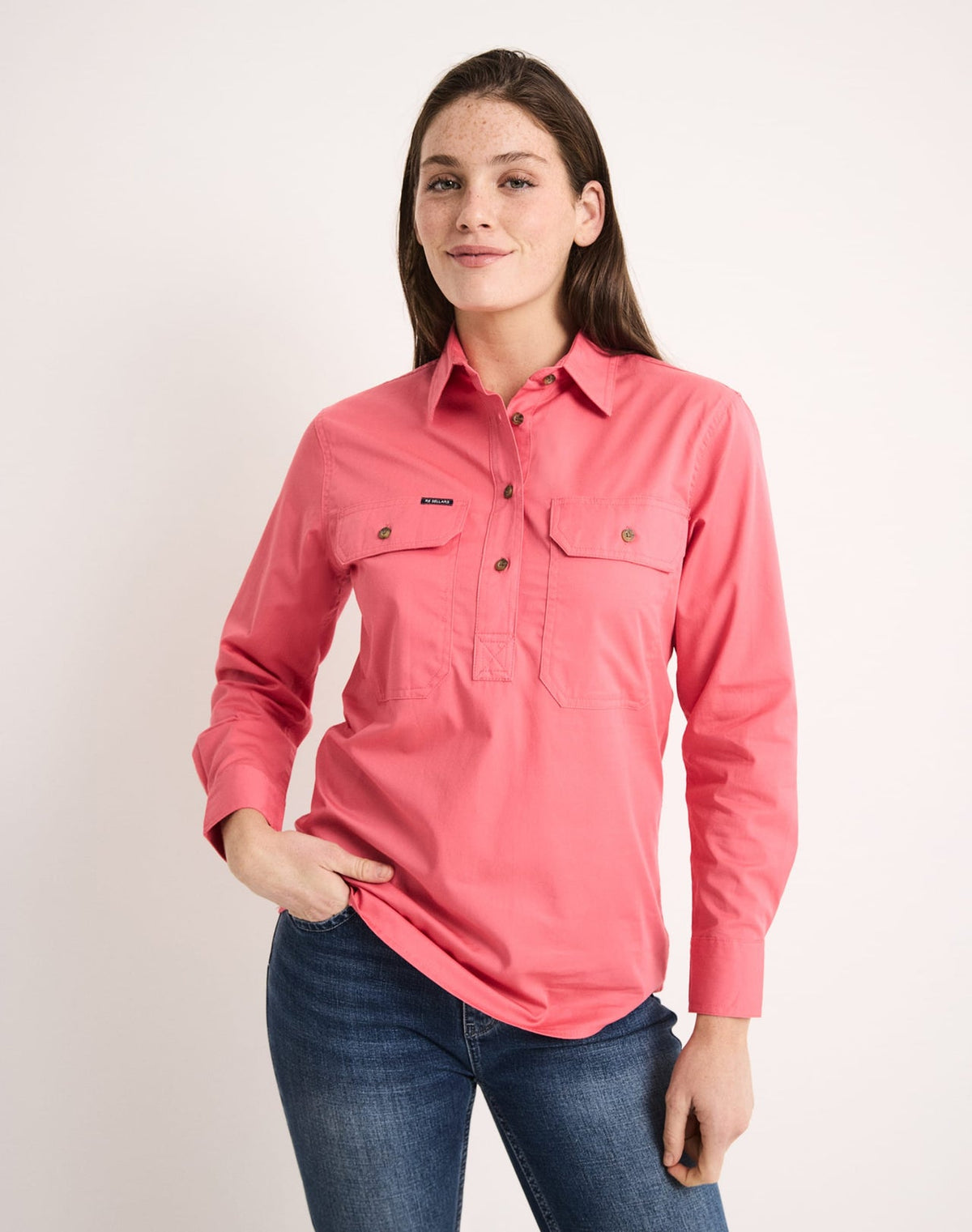 A woman modeling a coral pink long-sleeve button-up shirt with two chest flap pockets, paired with blue jeans, against a plain light background.